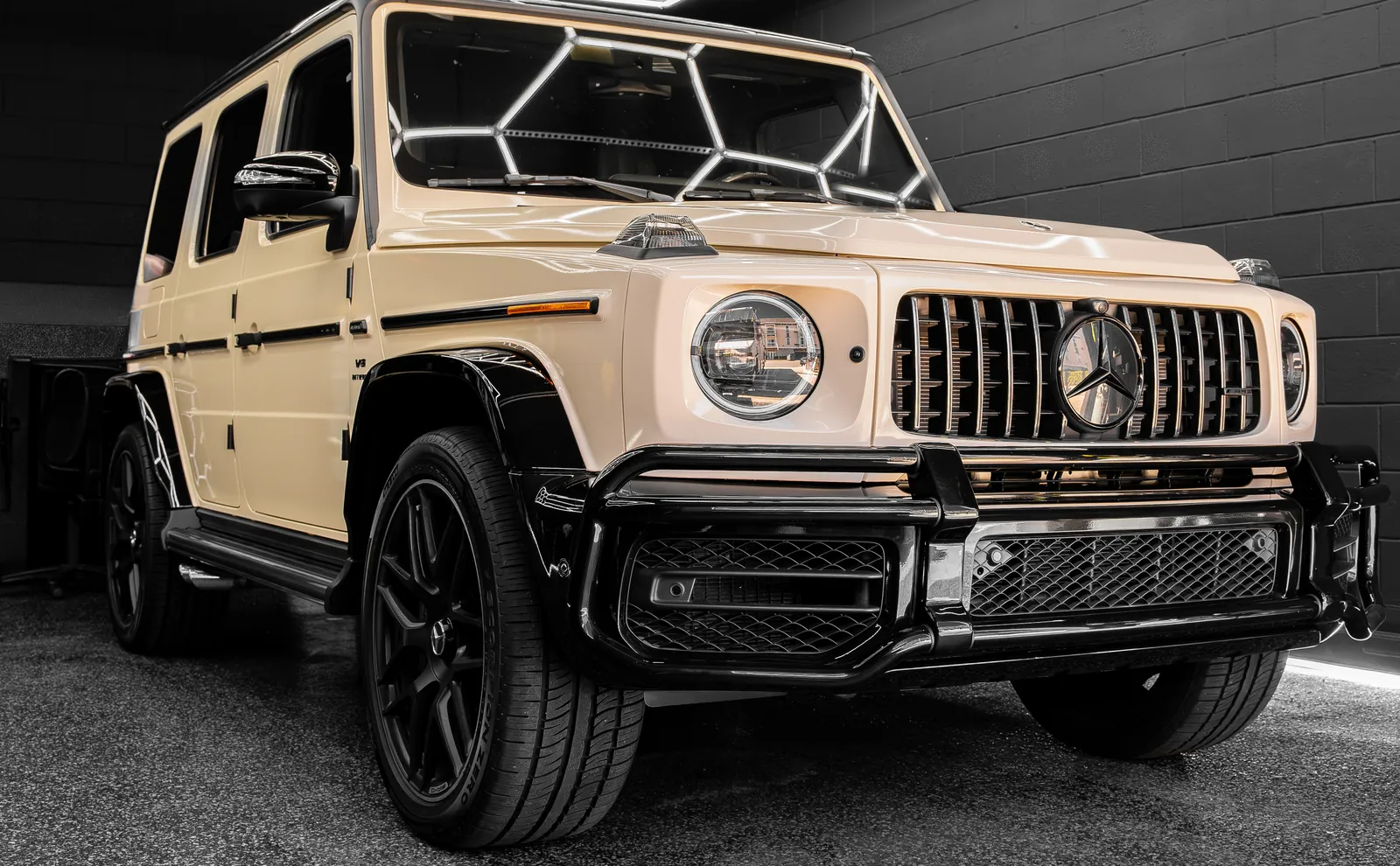 Beige Mercedes-AMG G63 with a vinyl wrap and gloss black accents, front three-quarter view in the shop bay