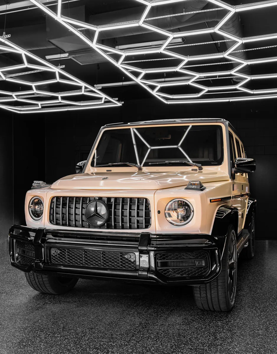 Front of a beige Mercedes-AMG G63 under the hex ceiling lights in the shop bay